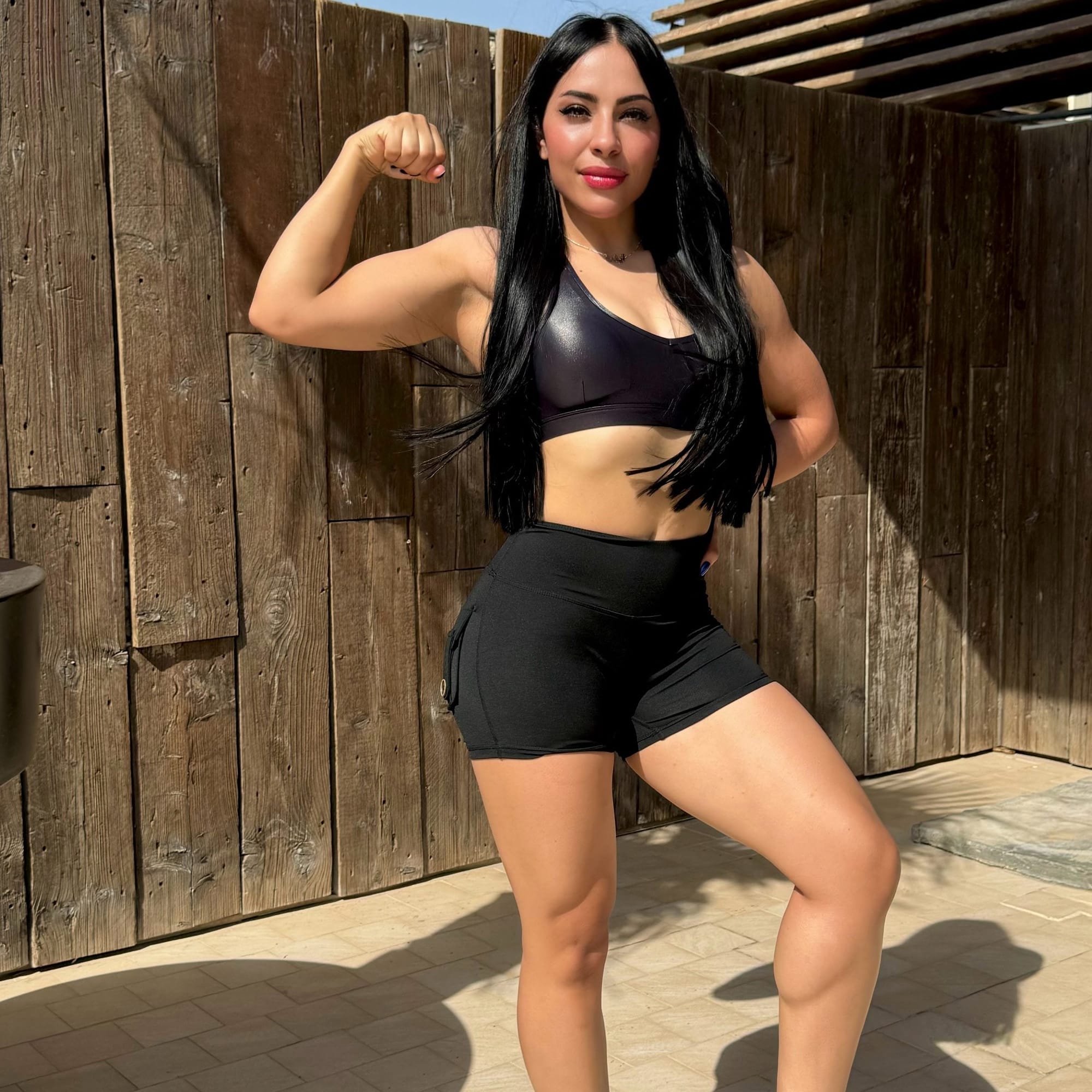 A woman with long black hair is wearing a black sports bra and shorts, striking a strong flexing pose. She is outdoors, standing on a tiled surface with a wooden fence in the background. The sunlight casts a clear shadow, highlighting her muscular physique.