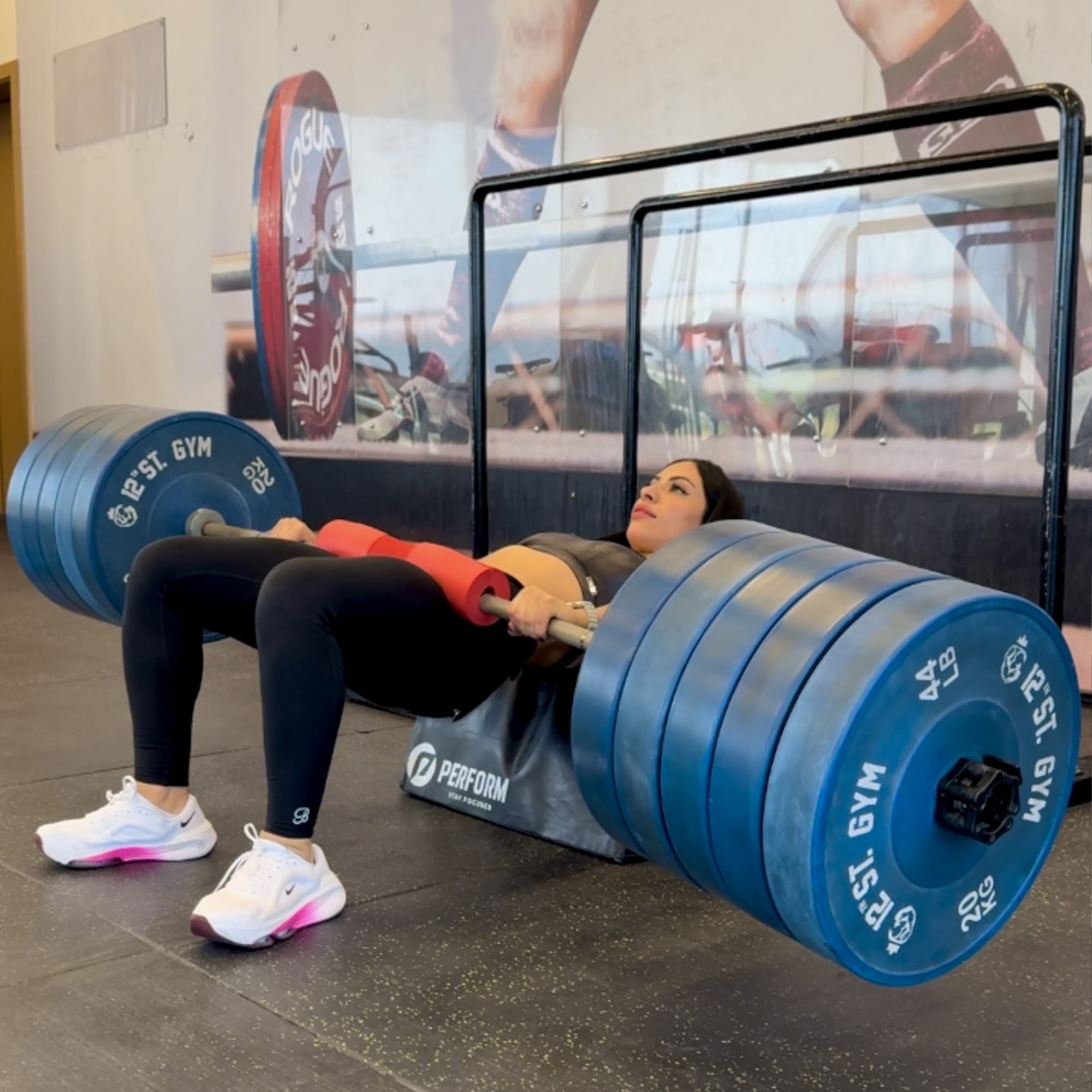 A woman performs a hip thrust exercise at the gym. She's lying on her back, using a barbell with multiple blue weight plates. She wears black leggings, a black top, and white sneakers. A large mirror and gym equipment are visible in the background.