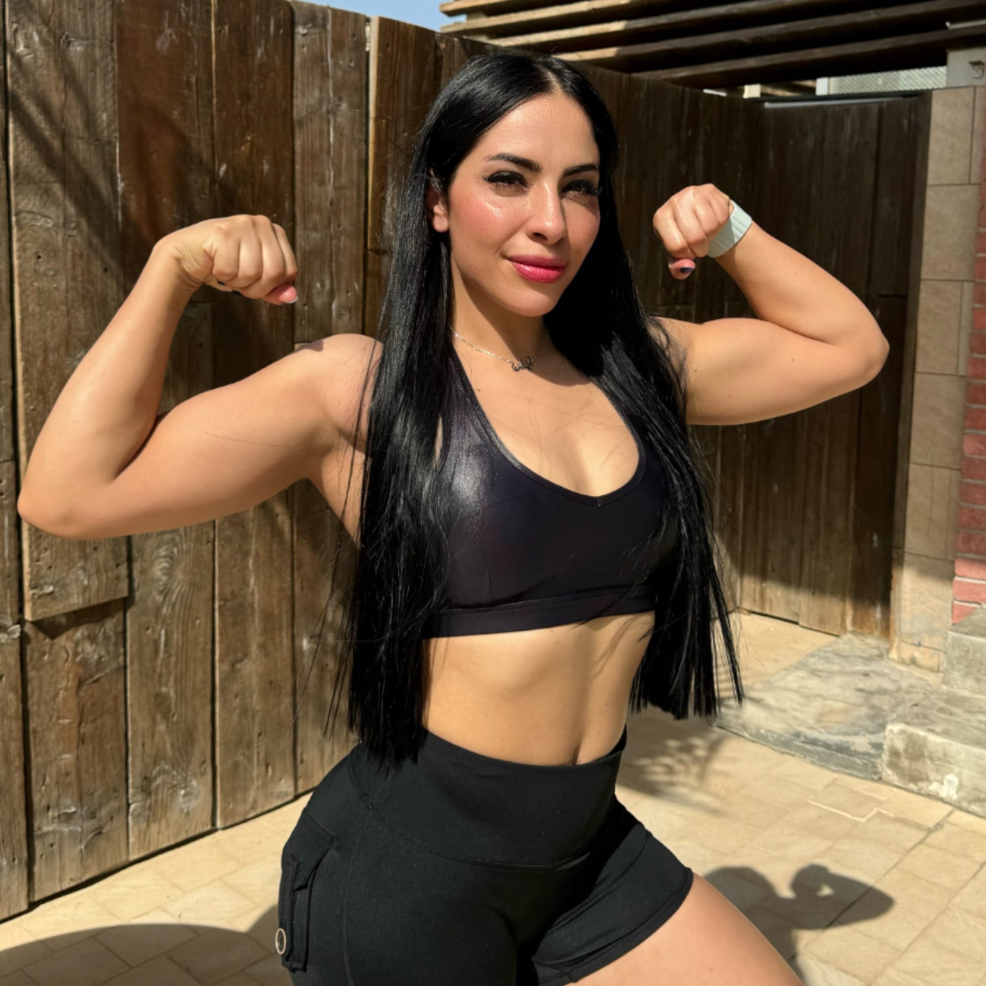A woman flexes her arm muscles while smiling confidently outdoors. She has long black hair and is wearing a black sports bra and black shorts. A wooden fence and part of a brick wall are visible in the background under bright sunlight.